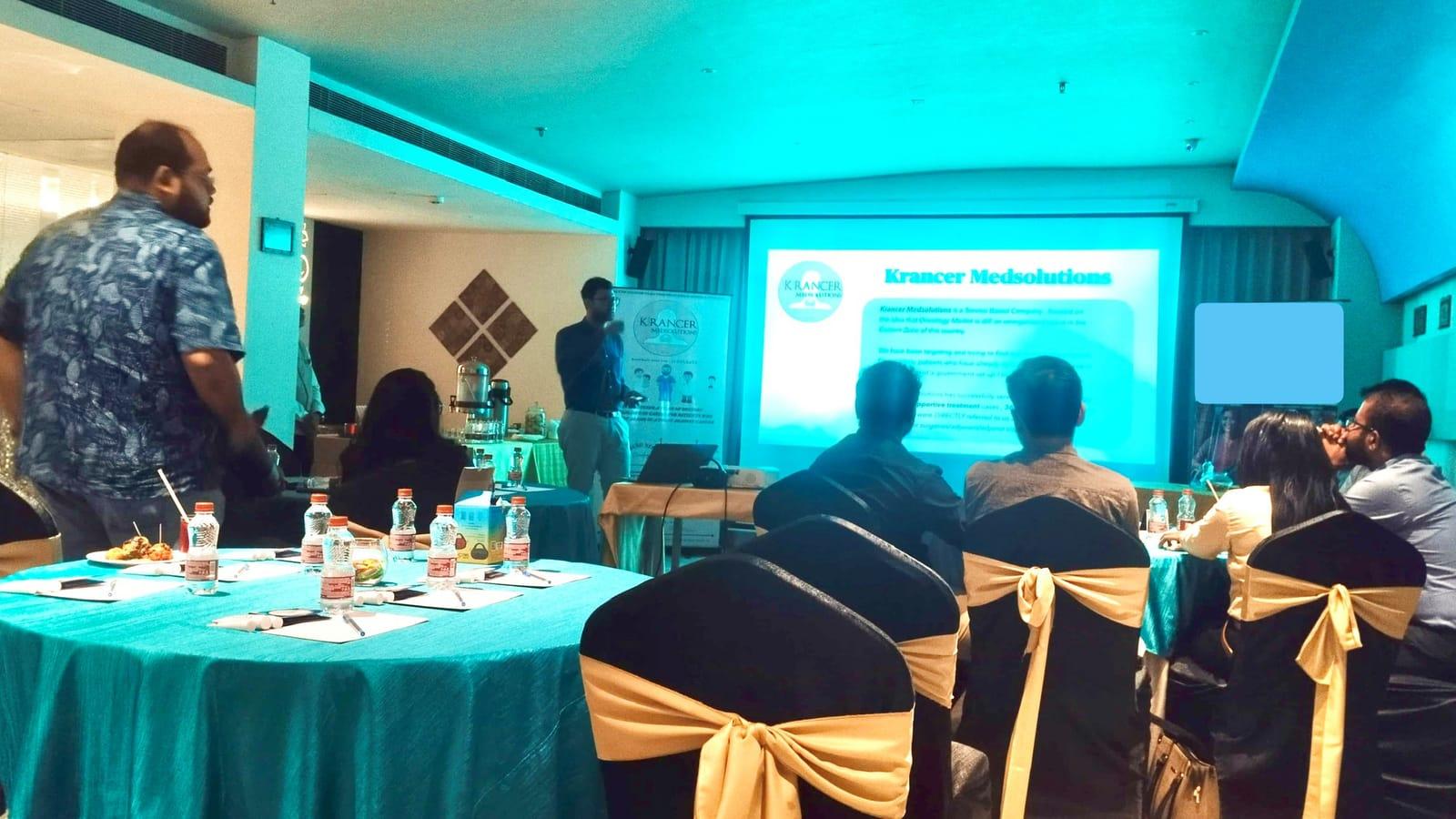 A Krancer doctor presenting to a seated audience, with a Krancer Medsolutions slide on the screen